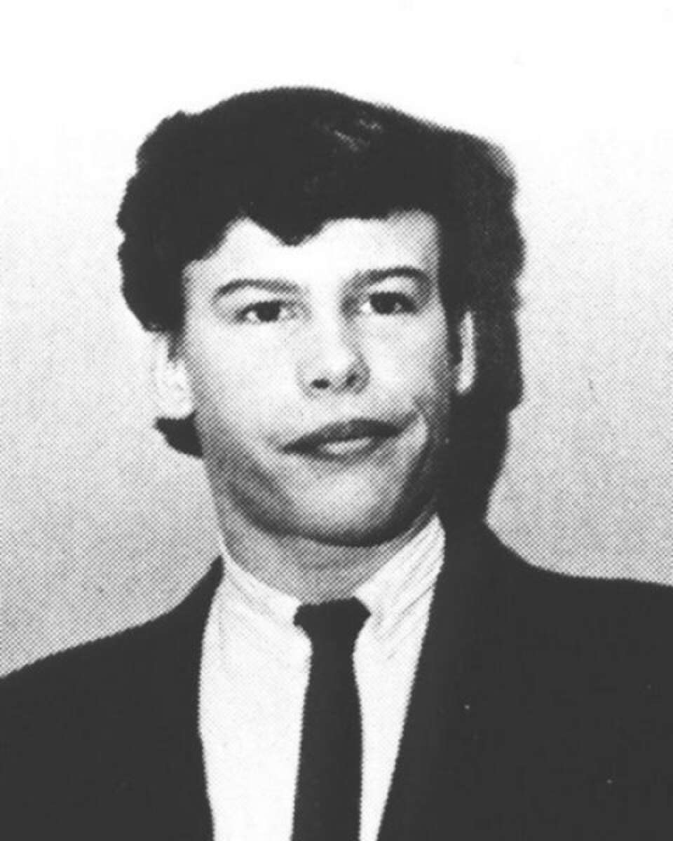 A black and white photo of a young person with short, dark hair wearing a suit, white shirt, and dark tie, looking slightly to the side with a neutral expression.