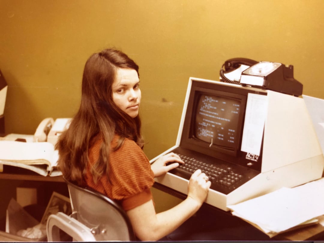 A young woman with long brown hair sits at a vintage computer terminal, typing. She looks over her shoulder towards the camera. Papers and an old rotary phone are on the desk. The room has olive green walls.