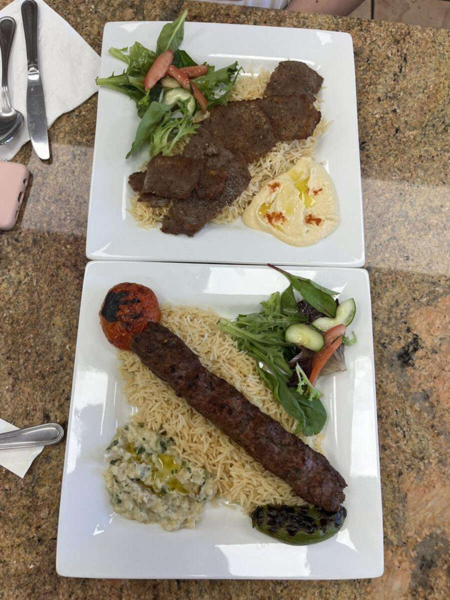 Two square plates with Middle Eastern food: one plate has sliced grilled meat over rice, salad, and hummus; the other has a long kebab, rice, grilled tomato, grilled pepper, salad, and a creamy dip. Both are on a granite tabletop.