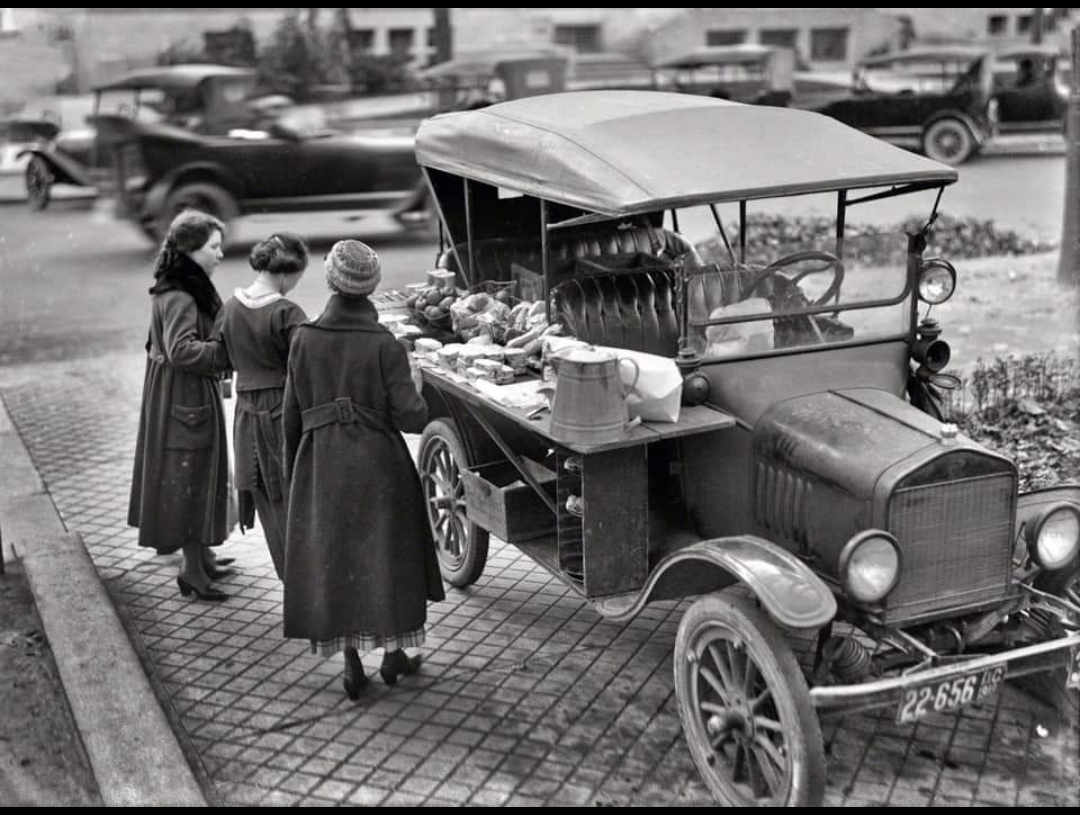 Three women stand at a vintage truck roadside stall selling produce. The black-and-white photo shows early 20th-century cars and people wearing coats, on a city sidewalk with buildings and cars in the background.