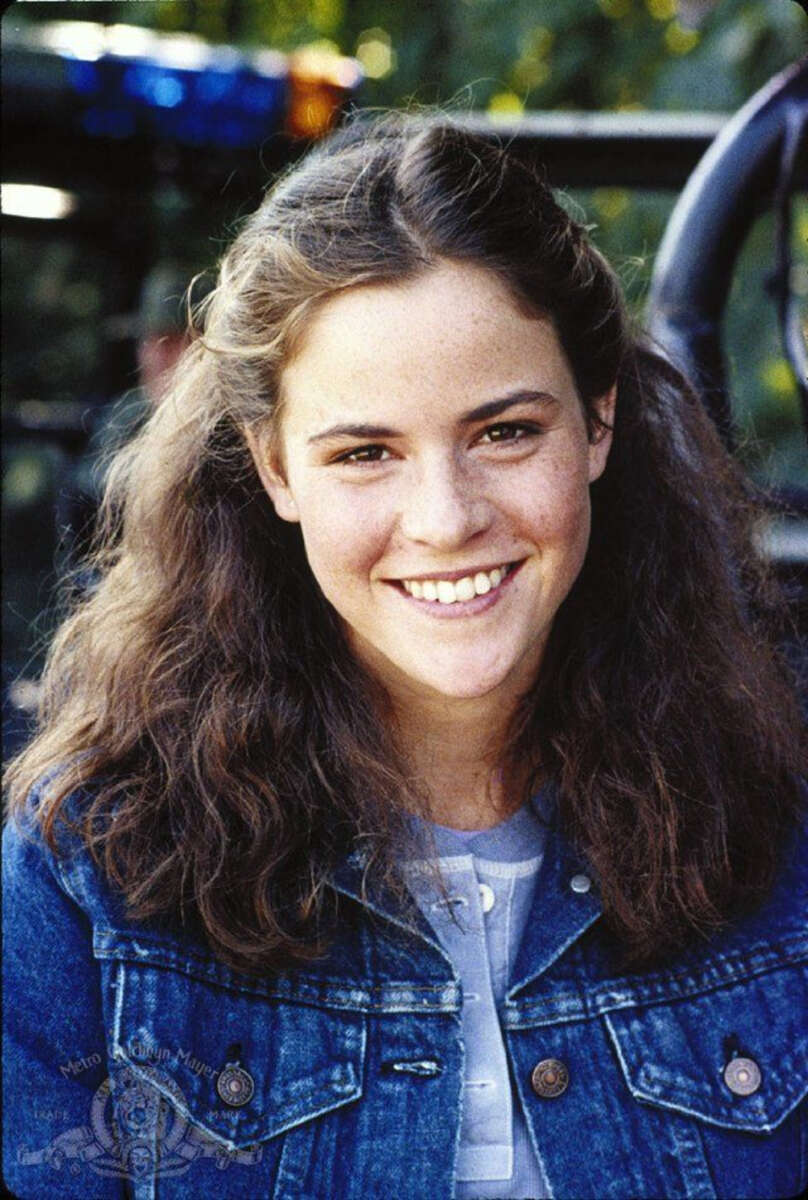 A young woman with wavy brown hair wearing a blue denim jacket and light-colored shirt smiles warmly at the camera. The background is slightly blurred with green foliage and some dark shapes.