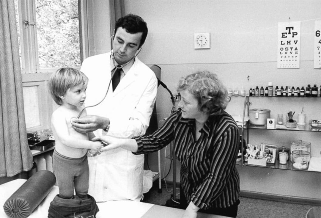 A doctor uses a stethoscope to examine a young child standing on an exam table while a woman, possibly the child’s mother, holds the child’s hand in a medical office. An eye chart and medical supplies are visible in the background.