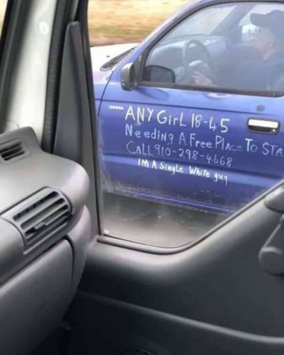 A blue car has a message written on its door in white marker, reading: "ANY girl 18-45 needing a free place to stay call 910-298-4668. I'm a single white guy." The photo is taken from inside another vehicle.