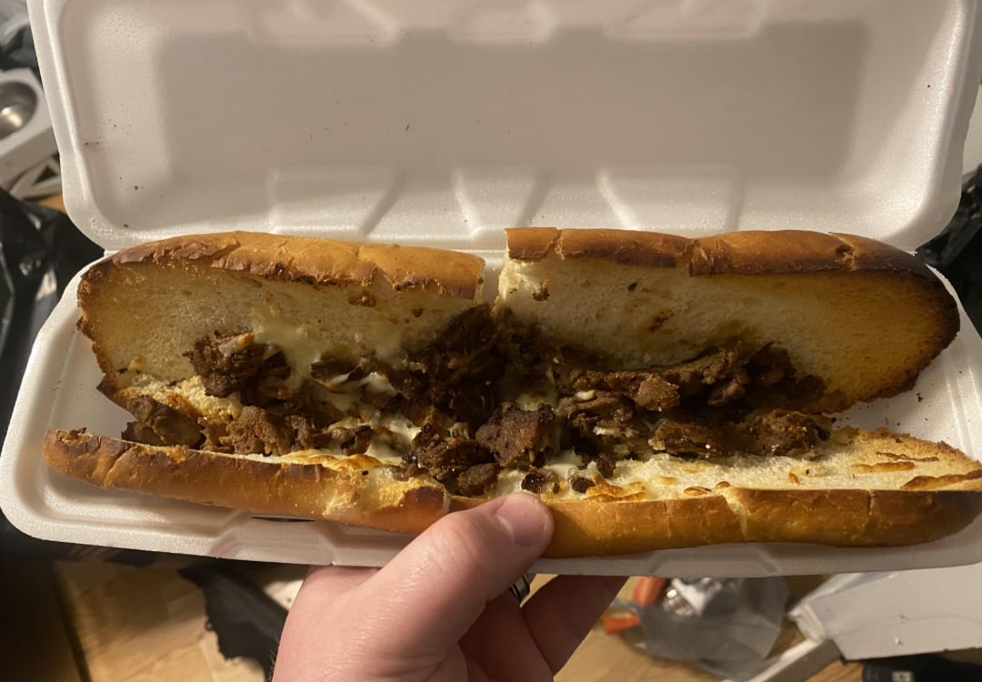 A hand holds an open foam takeout container with a sub sandwich inside, filled with chopped meat and melted cheese on a toasted bun.