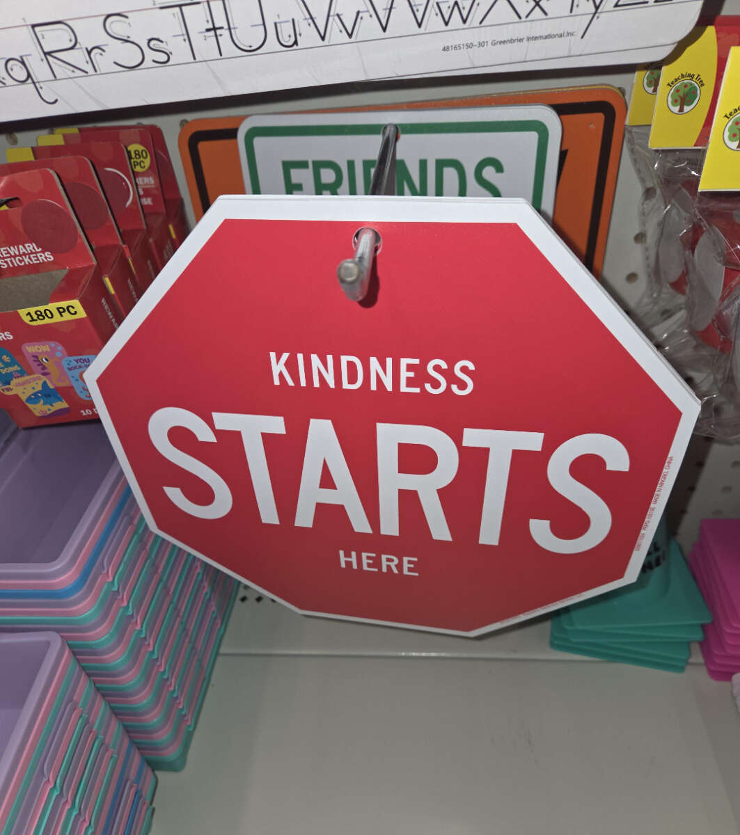 A red, octagon-shaped sign hanging in a store reads "KINDNESS STARTS HERE" in white letters. It resembles a stop sign. Colorful trays and other signs are displayed nearby.
