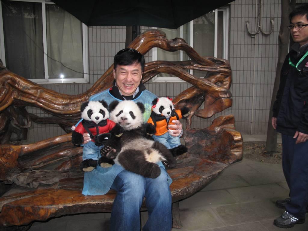 A smiling man wearing a blue apron sits on a wooden bench, holding a panda cub and two stuffed pandas dressed in colorful outfits, with two people standing nearby.