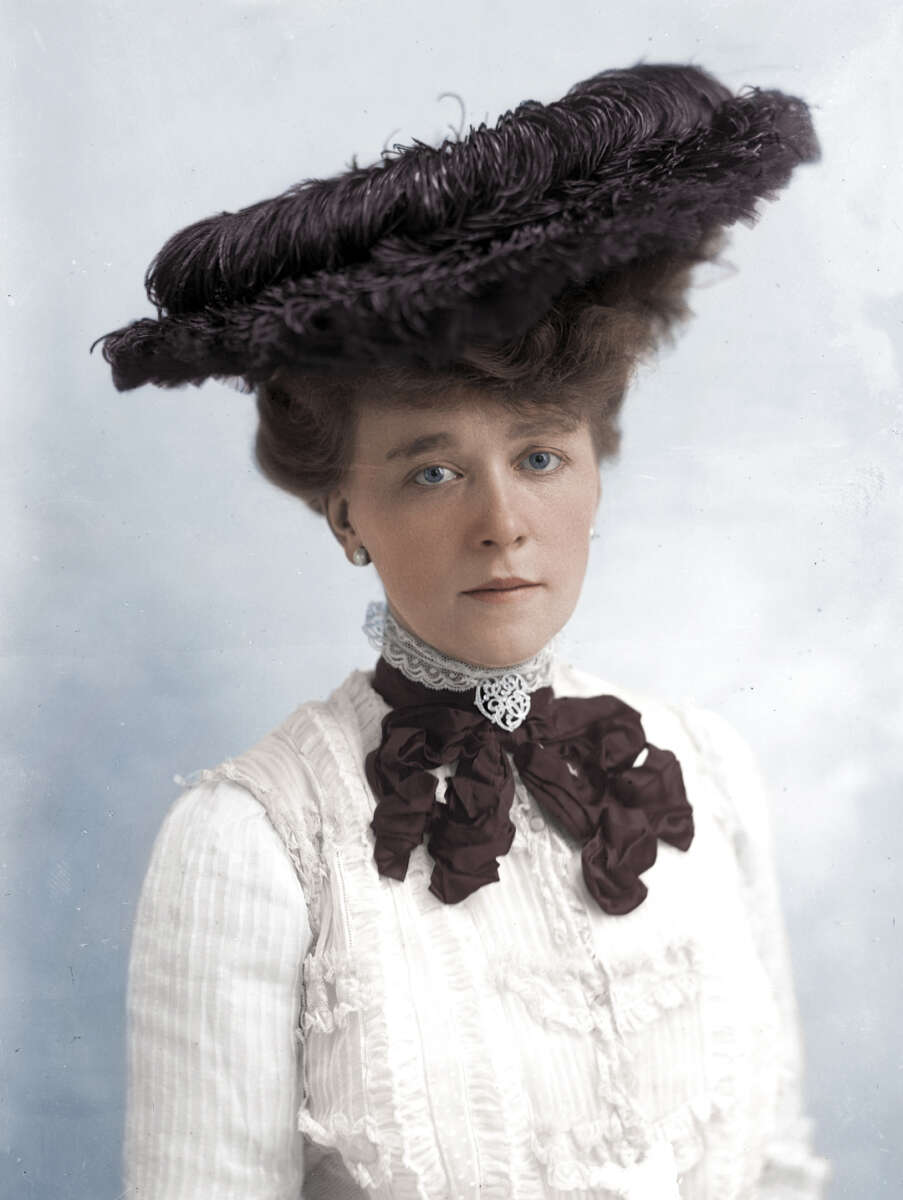 A woman with light-colored eyes and fair skin wears a large, dark feathered hat and a white high-collared blouse with lace and a dark ribbon bow at the neck, against a pale background.