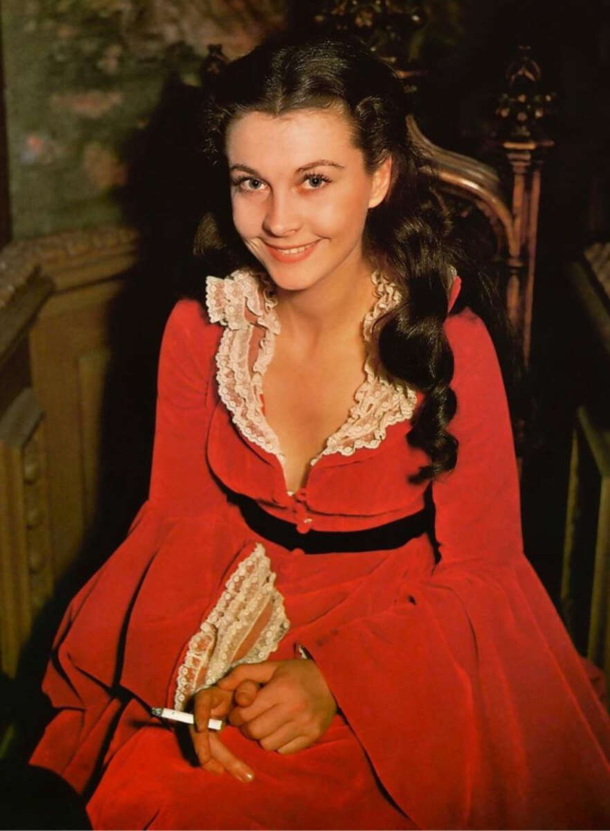 A woman with long dark hair in loose waves, wearing a bright red dress with white lace trim and a black belt, sits on a carved wooden chair and smiles at the camera while holding a cigarette in her hand.