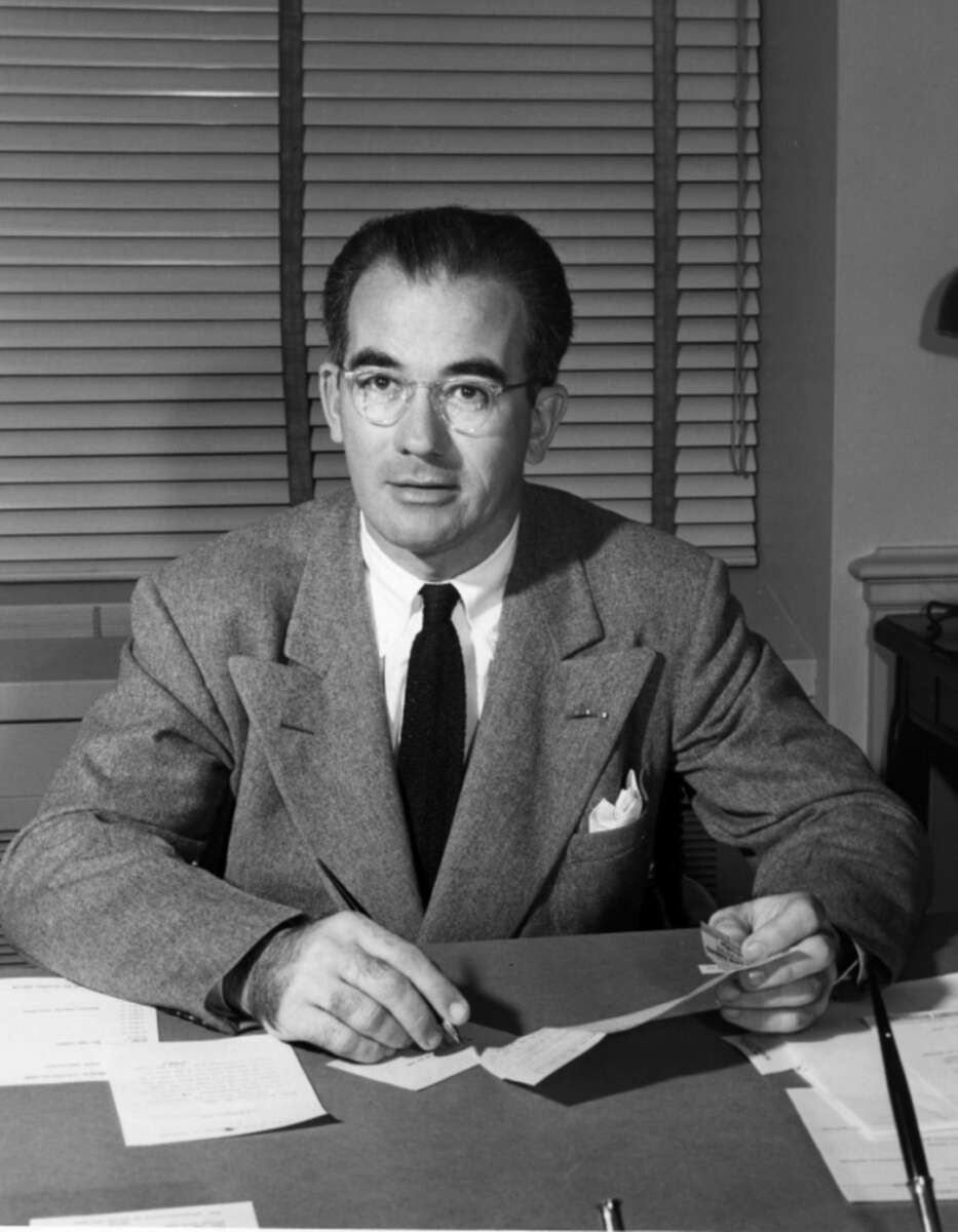 A man in a suit and tie sits at a desk with papers, holding a pen and looking toward the camera. Horizontal blinds cover the window behind him, and a lamp is partially visible on the right.