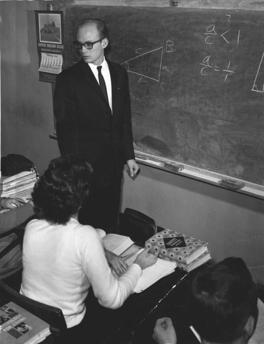 A teacher in a suit stands by a chalkboard with math formulas and triangle diagrams, while students sit at desks with open books and notebooks in a classroom.