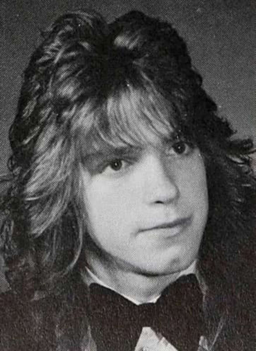 Black and white portrait of a young person with long, wavy hair, wearing a suit jacket, white shirt, and a large bow tie, facing slightly to the right with a neutral expression.
