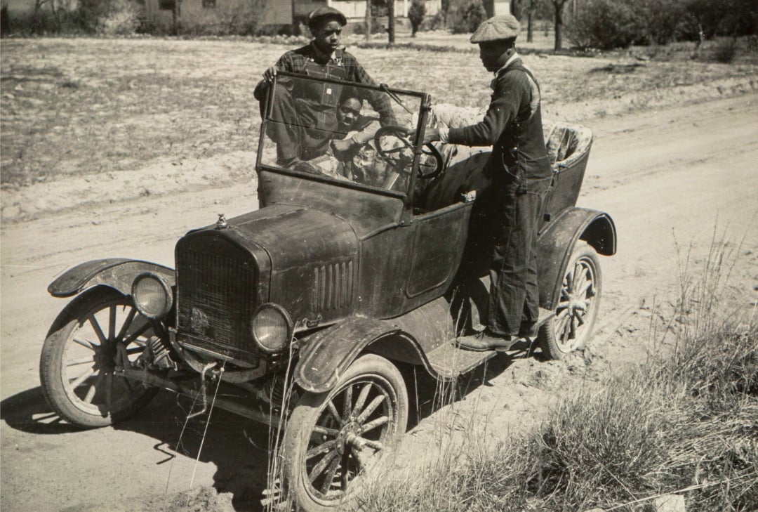 Two men with hats and jackets stand by or sit in an old, open-top car stuck in a dirt road, with grassy terrain and a house visible in the background.