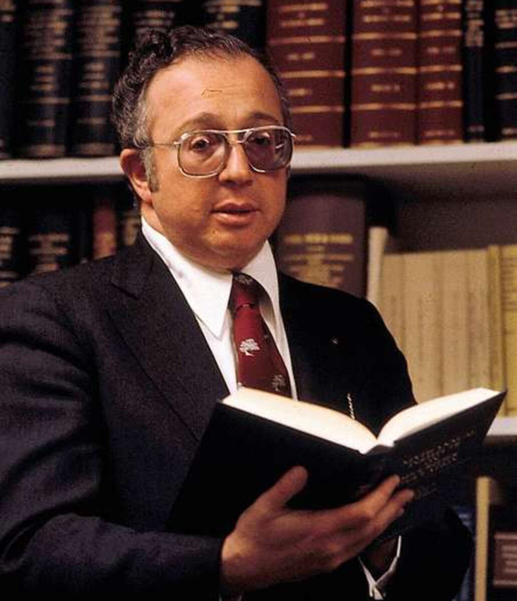 A man wearing glasses and a suit holds an open book in front of bookshelves filled with law or reference books. He faces the camera, appearing to be in a library or office setting.