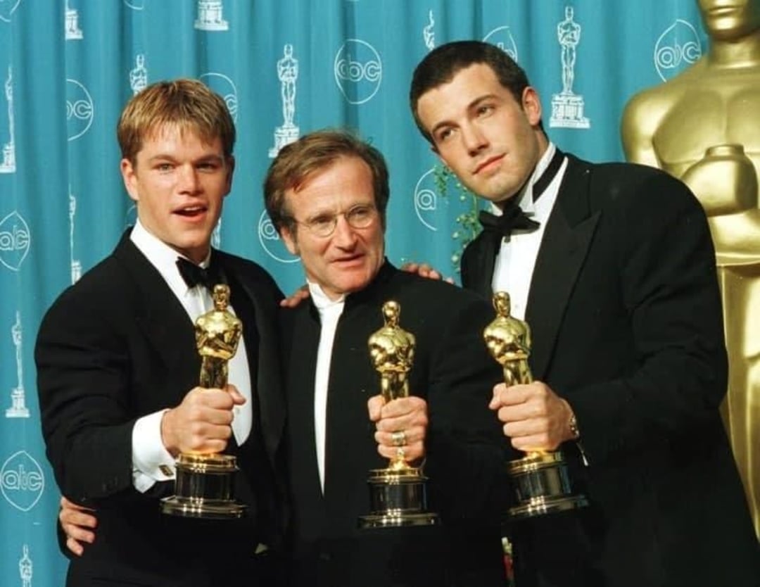 Three men in tuxedos are posing together, each holding an Oscar trophy, in front of an Oscar-themed backdrop at an awards ceremony.