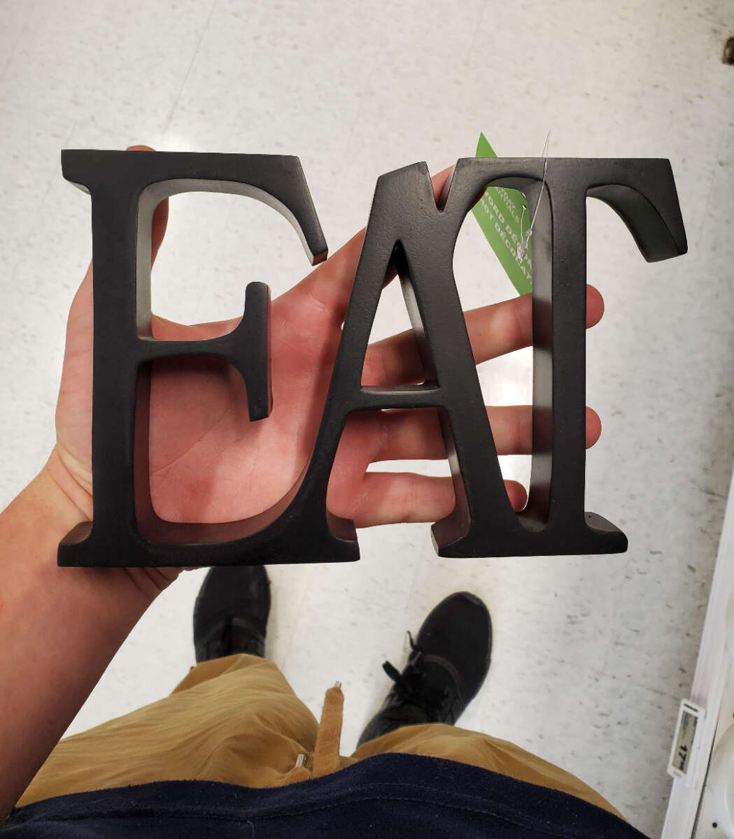 A person is holding a black decorative sign that spells out the word "EAT" in large capital letters. The background is a white tiled floor, and part of the person’s legs, dressed in tan pants and black shoes, is visible.