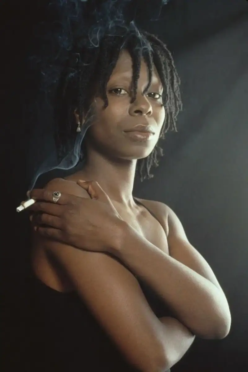 A woman with short, twisted dreadlocks stands against a dark background, crossing her arms and holding a lit cigarette. Smoke curls upward as she looks confidently at the camera, wearing a sleeveless black top.