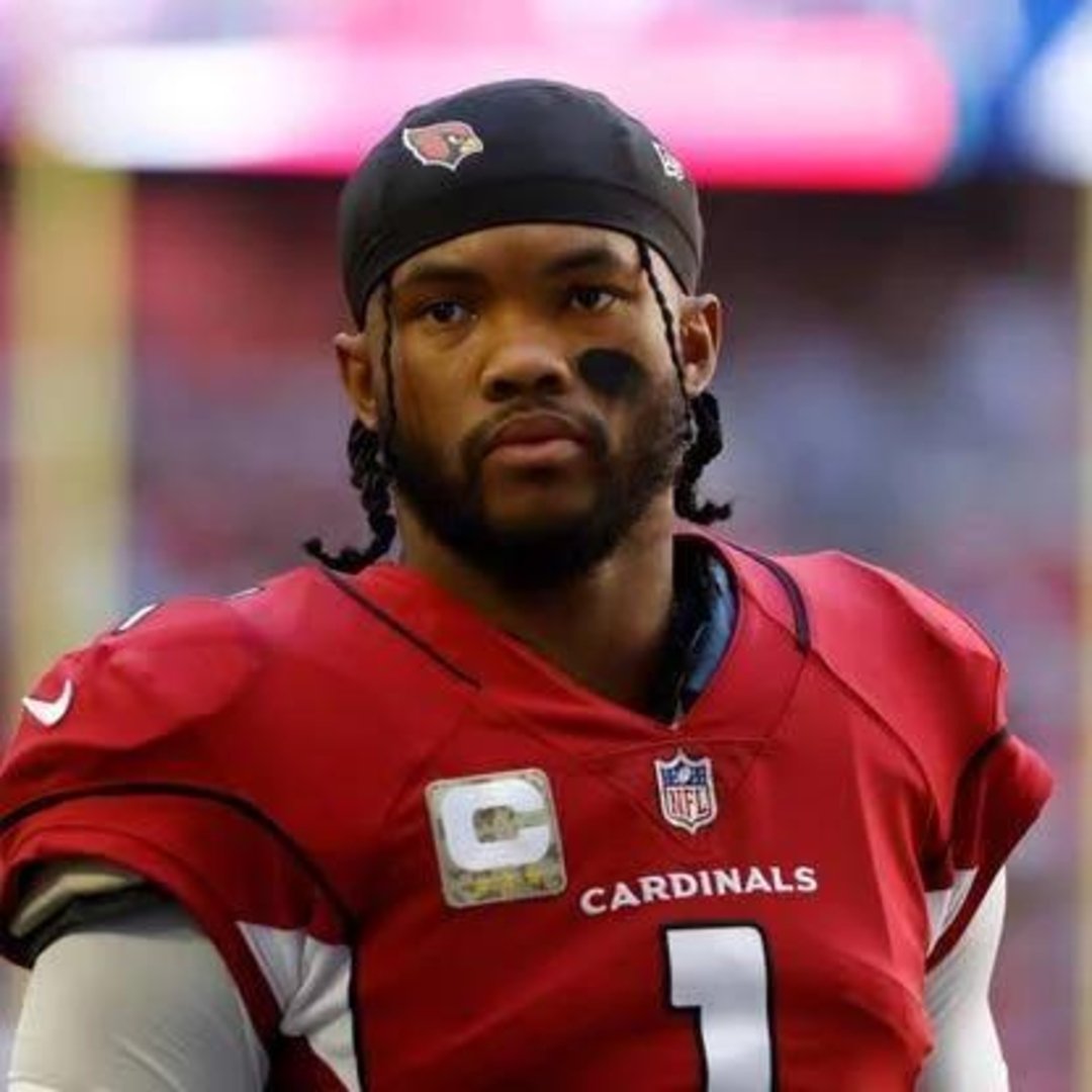 A football player in an Arizona Cardinals uniform with the number 1 on his jersey stands on the field, wearing a black headband and black eye paint under his eyes. The background is blurred.