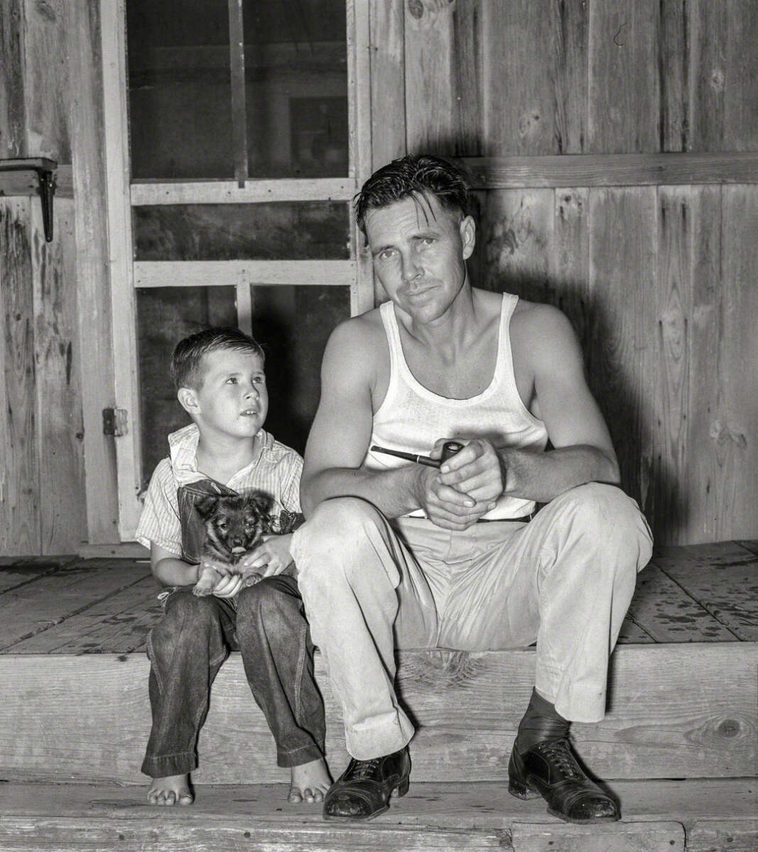 A man in a white tank top and light pants sits on a wooden porch next to a barefoot boy holding a cat. The man holds a pipe, and both look slightly to the side with serious expressions.