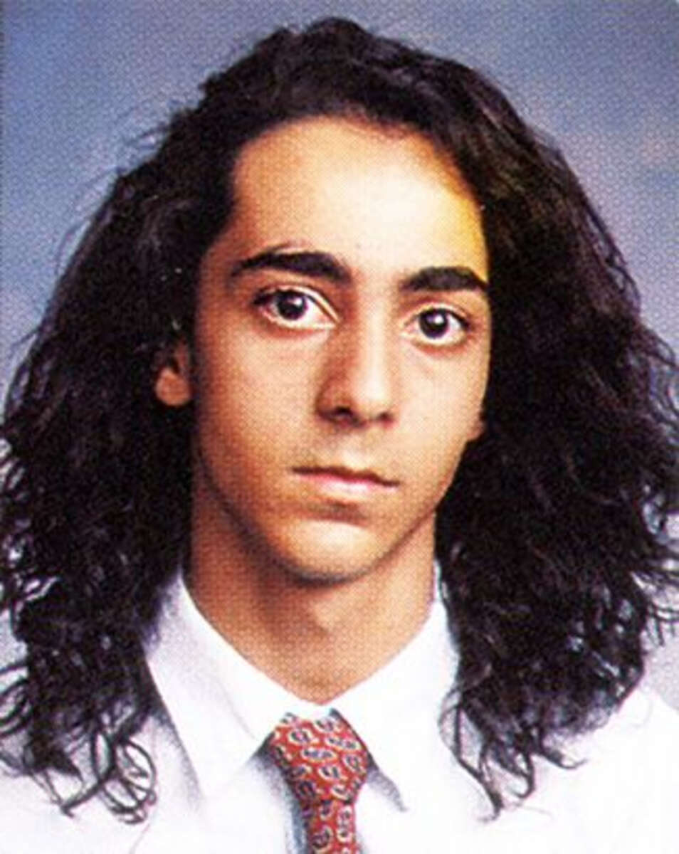 A young person with long, dark curly hair wearing a white shirt and a patterned tie poses for a formal portrait against a plain background.