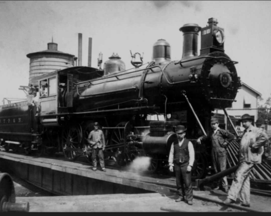 A black-and-white photo shows a vintage steam locomotive on a turntable, with four men in early 20th-century clothing standing nearby. A water tower and buildings are visible in the background.