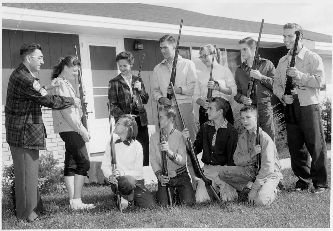 A black-and-white photo shows a group of young men and women, some kneeling and some standing, holding rifles outside a house, with an older man instructing them.