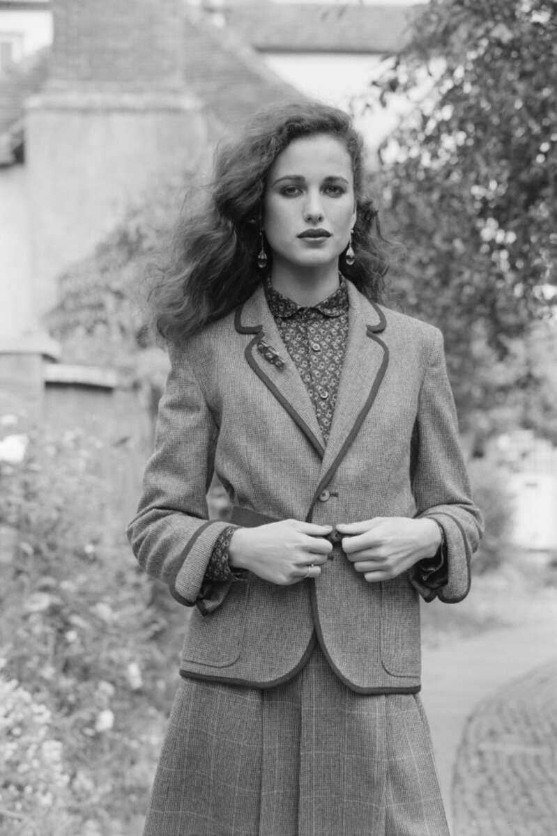 A woman with wavy hair stands outdoors, wearing a tailored blazer and patterned blouse. She looks confidently at the camera, hands holding the blazer. Blurred greenery and buildings appear in the background. Black and white photo.