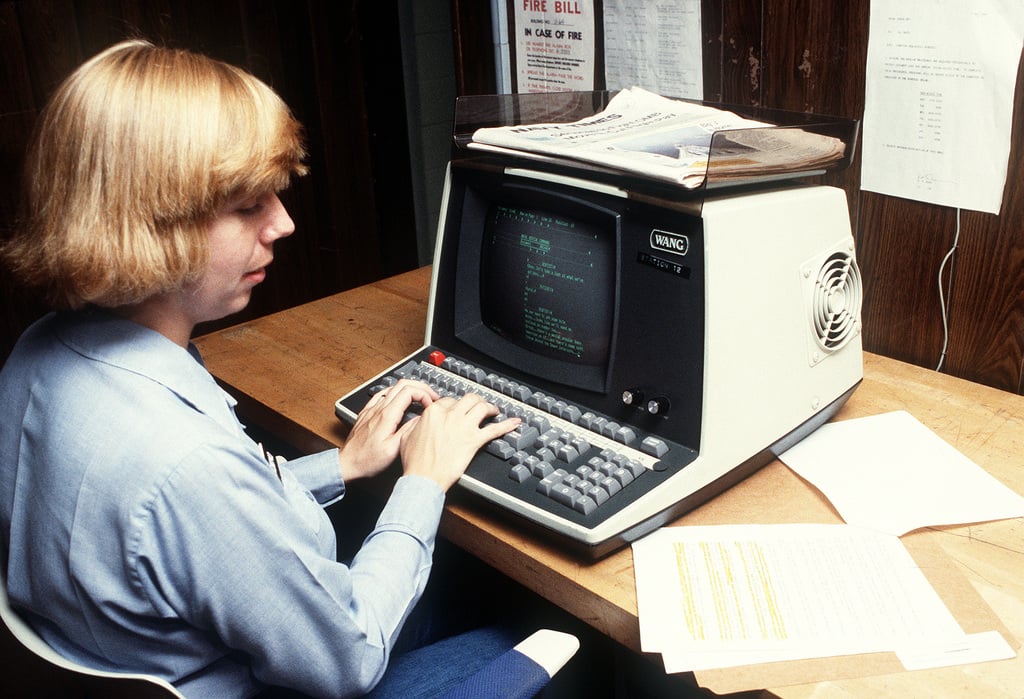 A person with blonde hair types on a vintage terminal computer in an office. Papers and a newspaper are on the desk, and documents are pinned to a wooden wall in the background.