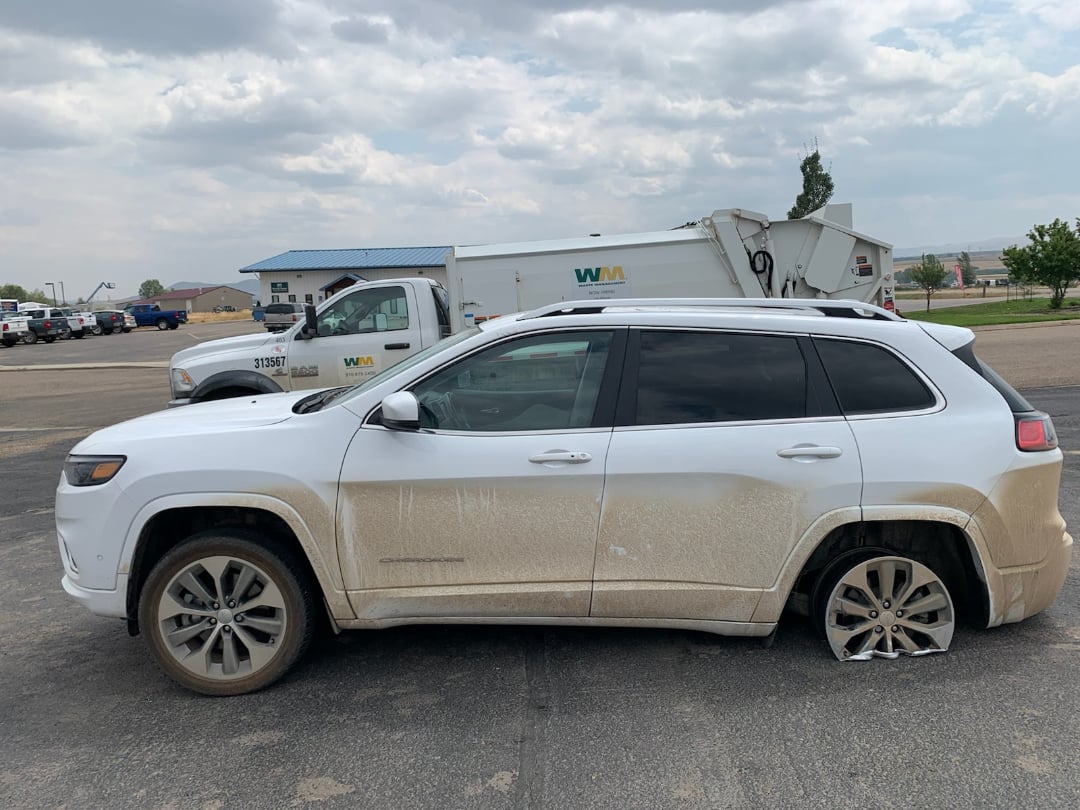 A white SUV with significant dirt on the doors is parked on pavement with a flat rear tire. In the background, there is a white Waste Management truck and a building under a cloudy sky.