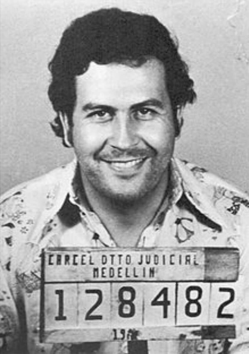 A man with dark curly hair smiles in a black-and-white mugshot photo, holding a sign that reads, "CARCEL OTTO JUDICIAL MEDELLIN 128482.