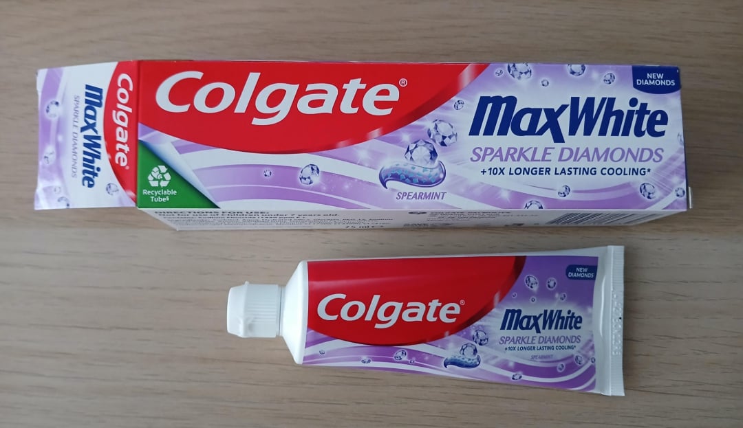 A box and tube of Colgate MaxWhite Sparkle Diamonds toothpaste with a purple and red design, featuring spearmint flavor and text highlighting longer-lasting cooling. Both items are on a light wooden surface.