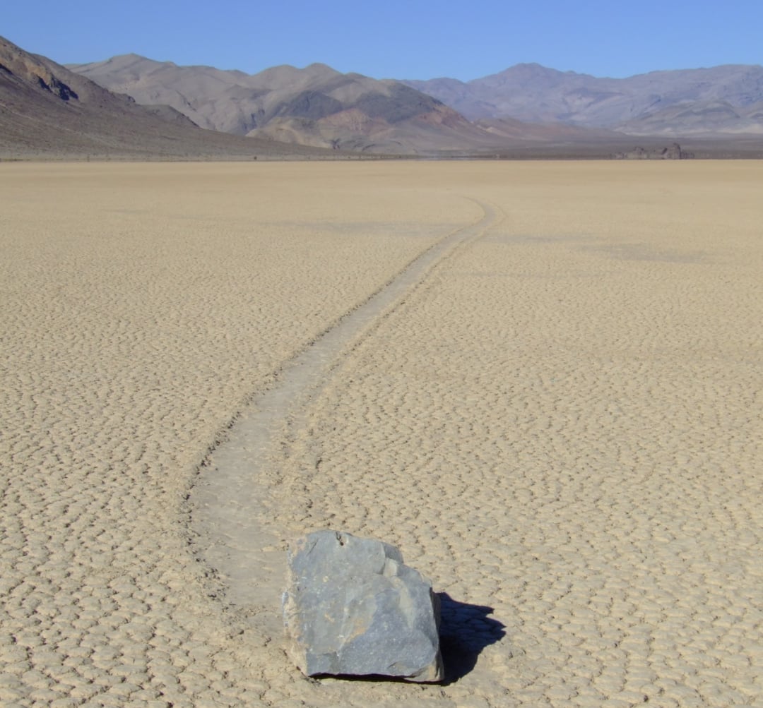 A large rock sits on cracked, dry desert ground, with a long, curved trail behind it, suggesting the rock has moved across the surface. Mountain ranges are visible in the distance under a clear blue sky.