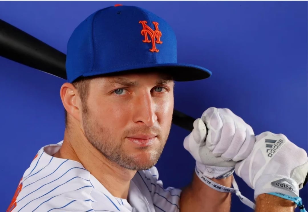 A baseball player in a blue New York Mets cap and striped jersey holds a bat over his shoulder, wearing white Adidas gloves, posed against a solid blue background.