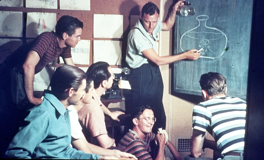 Six men, some in striped shirts, gather around a chalkboard as one man draws a scientific diagram of a flask with circles inside, possibly explaining a concept. Papers are pinned on the wall behind them.