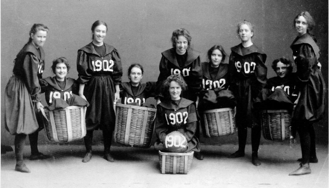 A black and white photo of nine women in uniforms marked "1902" posing with large baskets, likely an early women’s basketball team, arranged in two rows with a neutral studio backdrop.