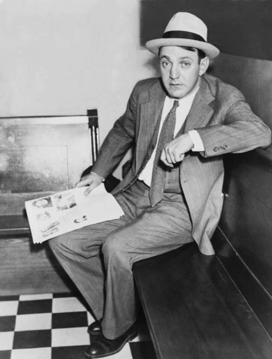 A man in a suit and tie, wearing a light-colored fedora, sits on a wooden bench holding a newspaper. He looks at the camera with a neutral expression. The floor is checkered black and white.