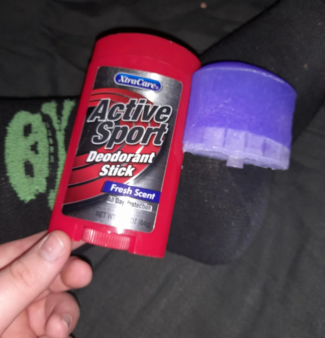 A hand holds a red XtraCare Active Sport deodorant stick near a leg with a purple plastic cap balanced on the ankle. The person is wearing black socks with a green smiley face design.