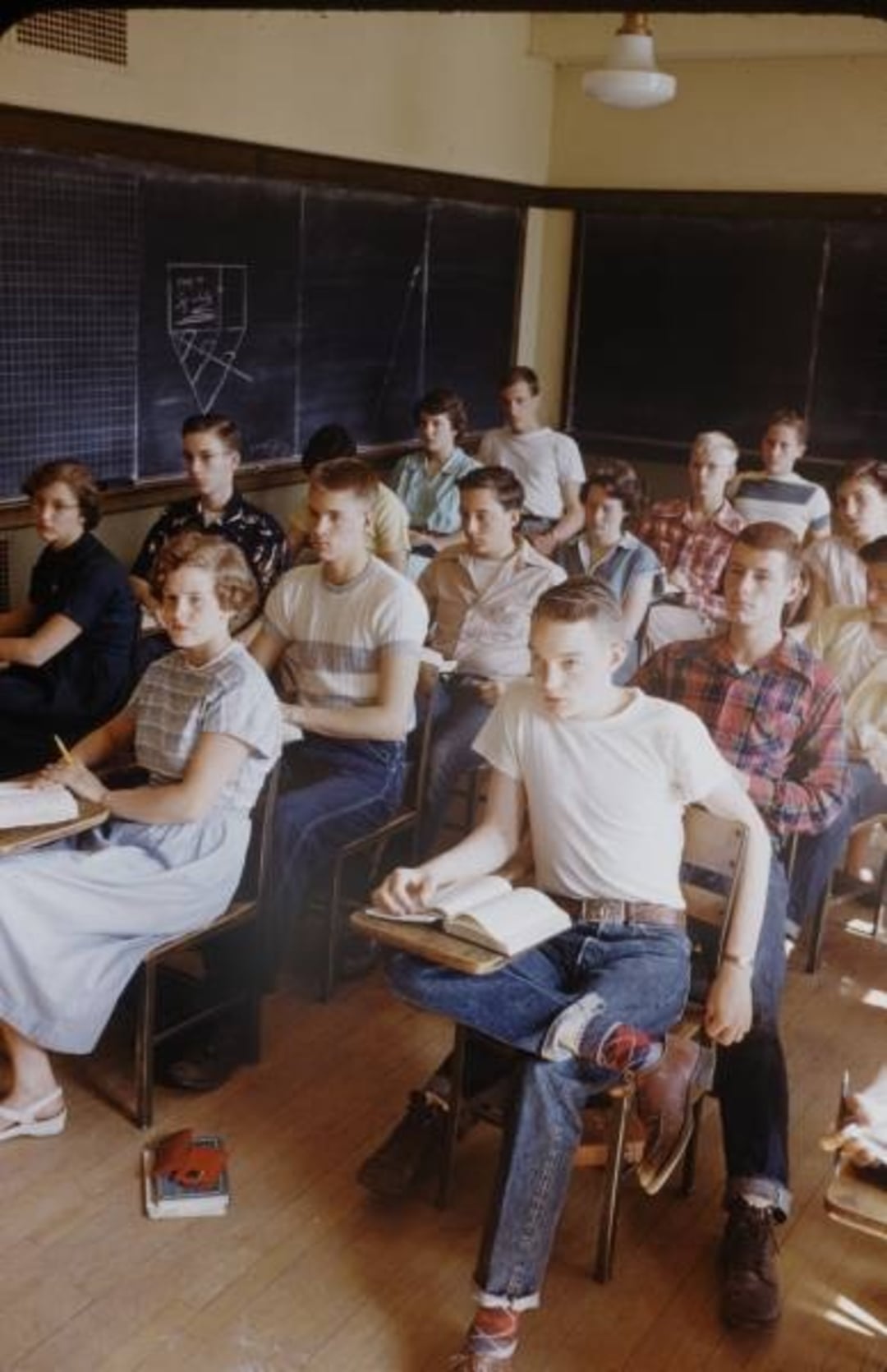 A group of teenagers sit attentively in a classroom, facing forward with notebooks and textbooks on their desks. The room has wooden floors, blackboards, and a chart drawn with chalk. The students appear to be listening to a lesson.