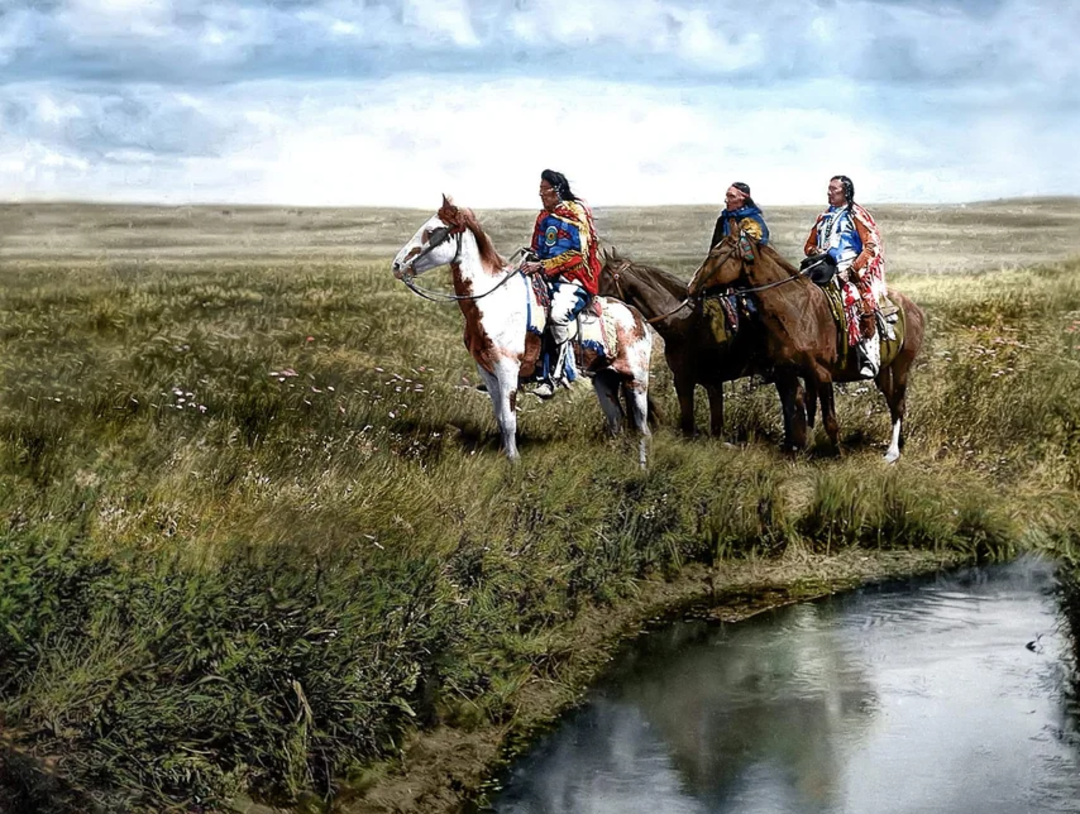 Three Indigenous people in traditional clothing sit on horses in a grassy field near a stream, looking into the distance under a partly cloudy sky.