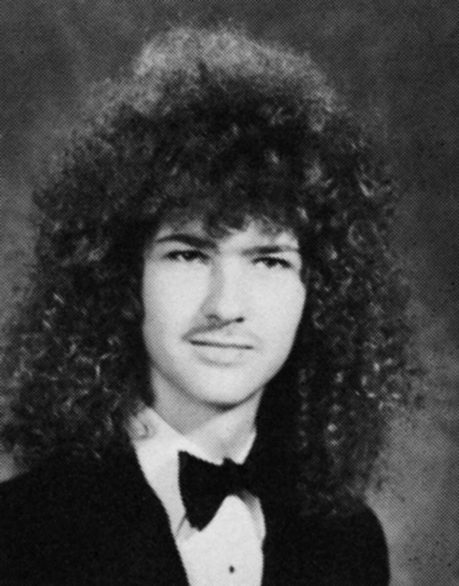 Black and white portrait of a young person with curly, voluminous hair, wearing a suit jacket, white shirt, and black bow tie, posing in front of a mottled studio backdrop.