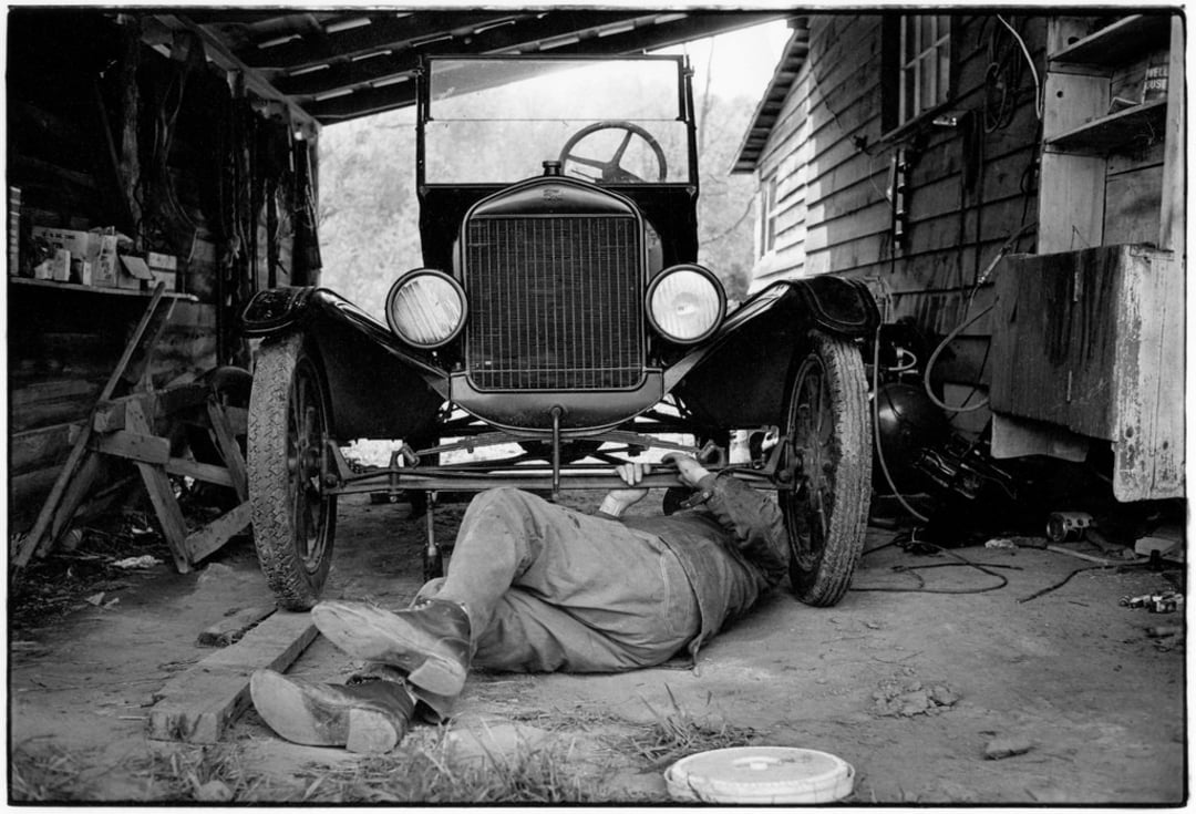 A person lies on the ground under a vintage car in a rustic garage, working on the vehicle's underside. Tools and equipment are scattered around the wooden structure, creating a classic, old-fashioned workshop atmosphere.