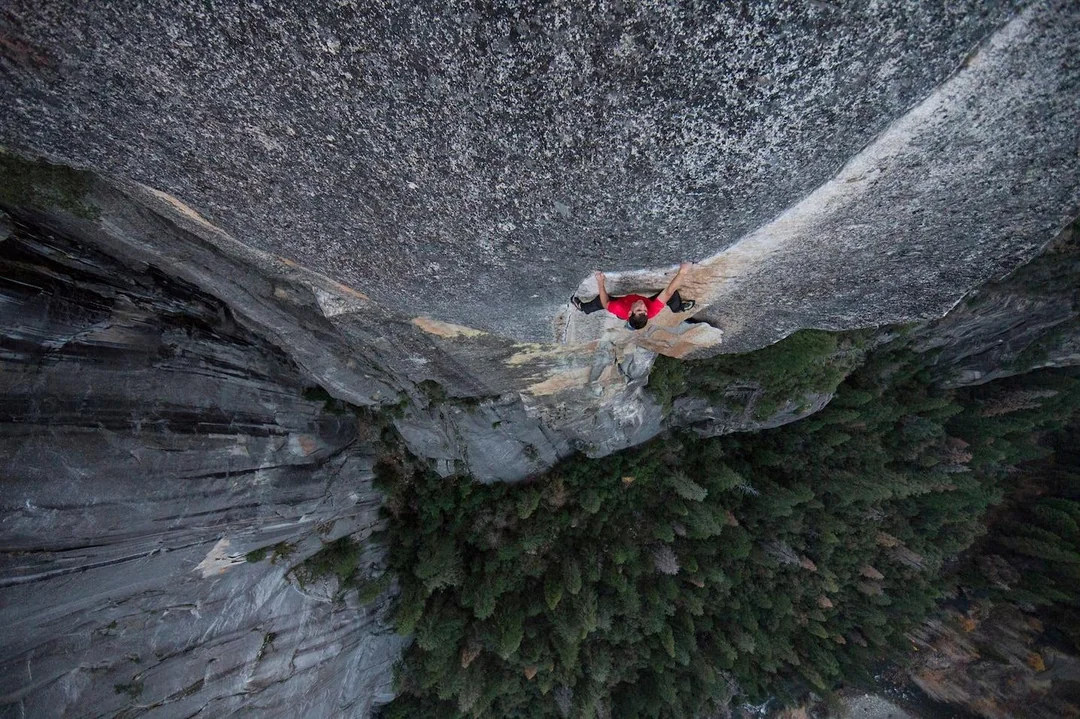 A rock climber in a red shirt ascends a sheer cliff face high above a dense forest, gripping the rock with both hands and feet, demonstrating skill and focus in a vertical landscape.