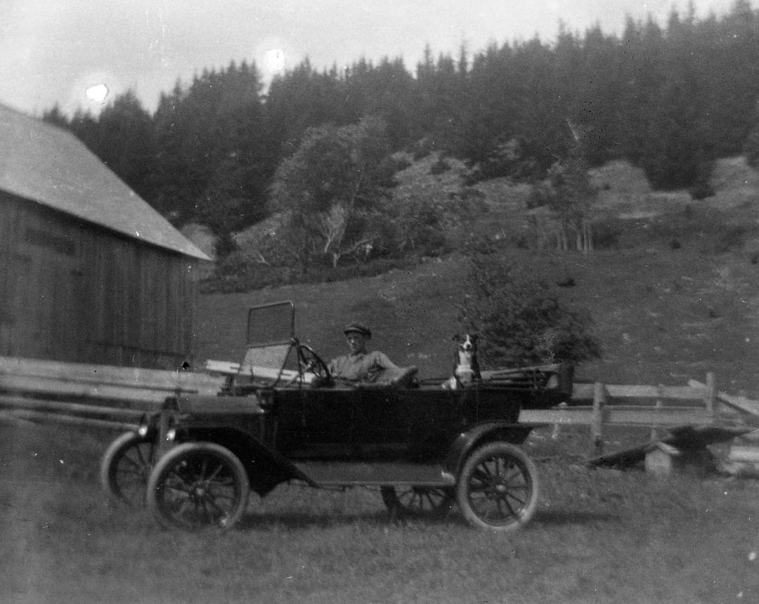 A black-and-white photo of a person sitting in an old-fashioned car parked on grass; a dog sits beside them. A wooden barn and a hillside with trees appear in the background.