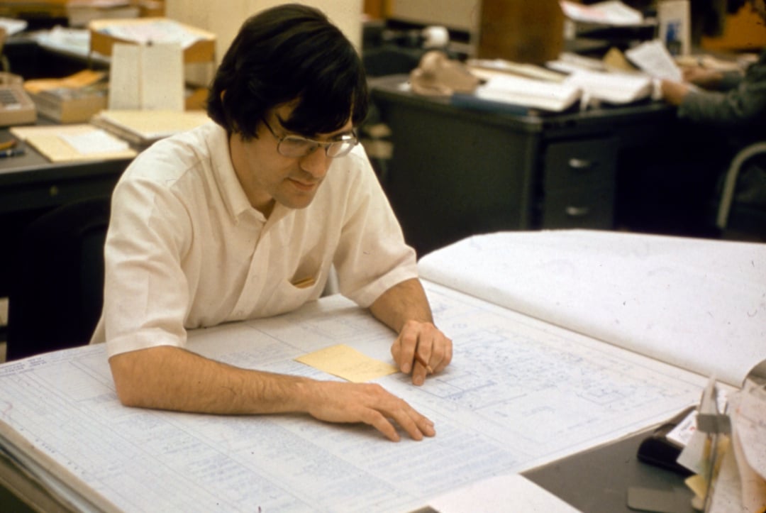 A man with glasses and dark hair examines large blueprints on a drafting table in an office, holding a pencil and a small notepad. Files, papers, and desks are visible in the background.