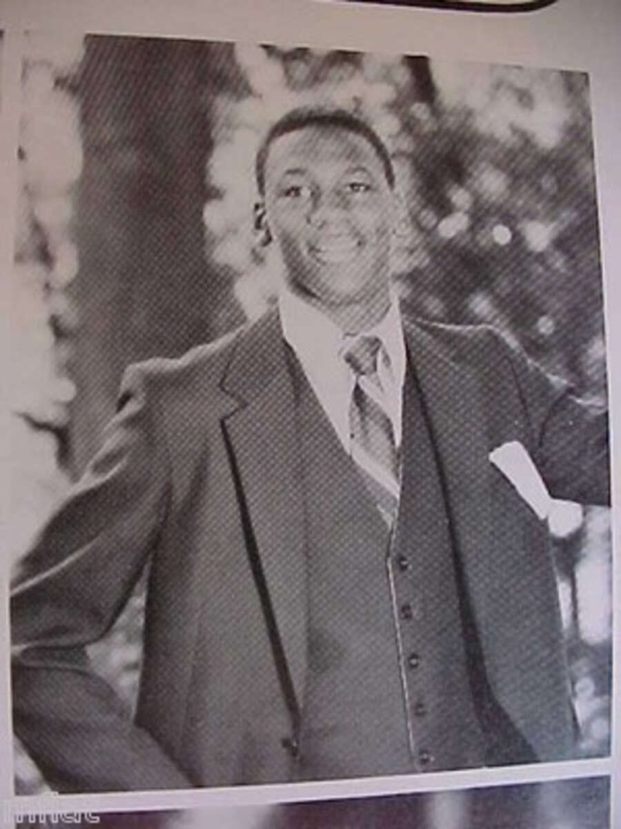 A young man in a suit and tie poses outdoors, smiling at the camera. The image is black and white, with trees blurred in the background.