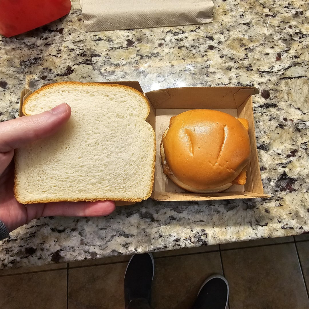 A hand holds a slice of white bread next to a boxed cheeseburger on a granite countertop. Part of a napkin, a red item, and a pair of feet on a tiled floor are also visible.