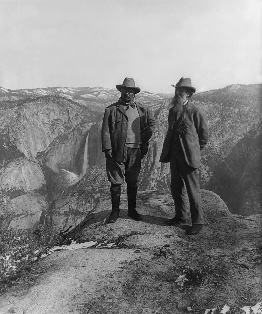 Two men in early 20th-century outdoor attire stand on a rocky cliff overlooking a deep valley and distant waterfall with forested mountains in the background.