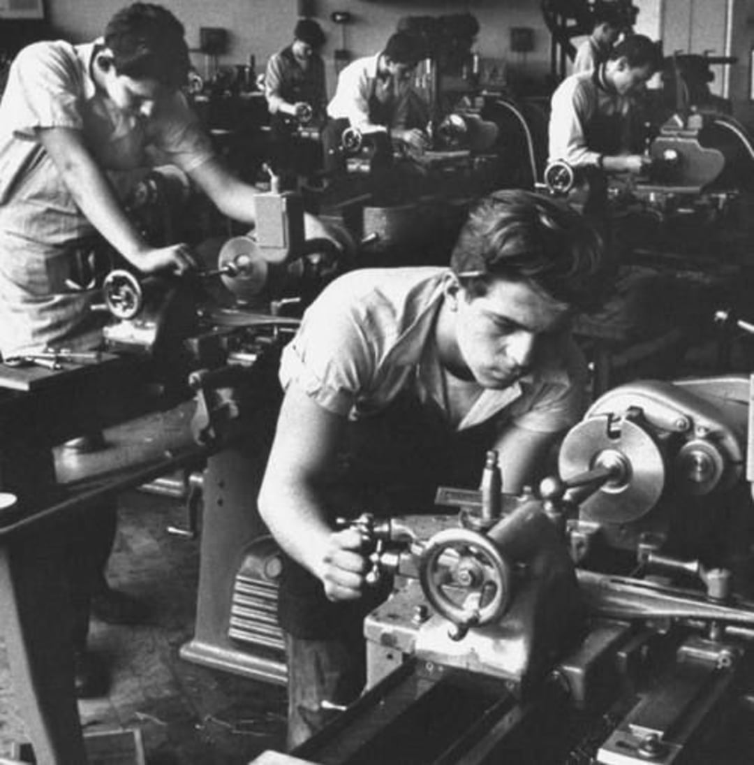 Several young men work intently on metal lathes in a workshop, wearing overalls and surrounded by machinery and tools, suggesting a hands-on technical or engineering training environment.