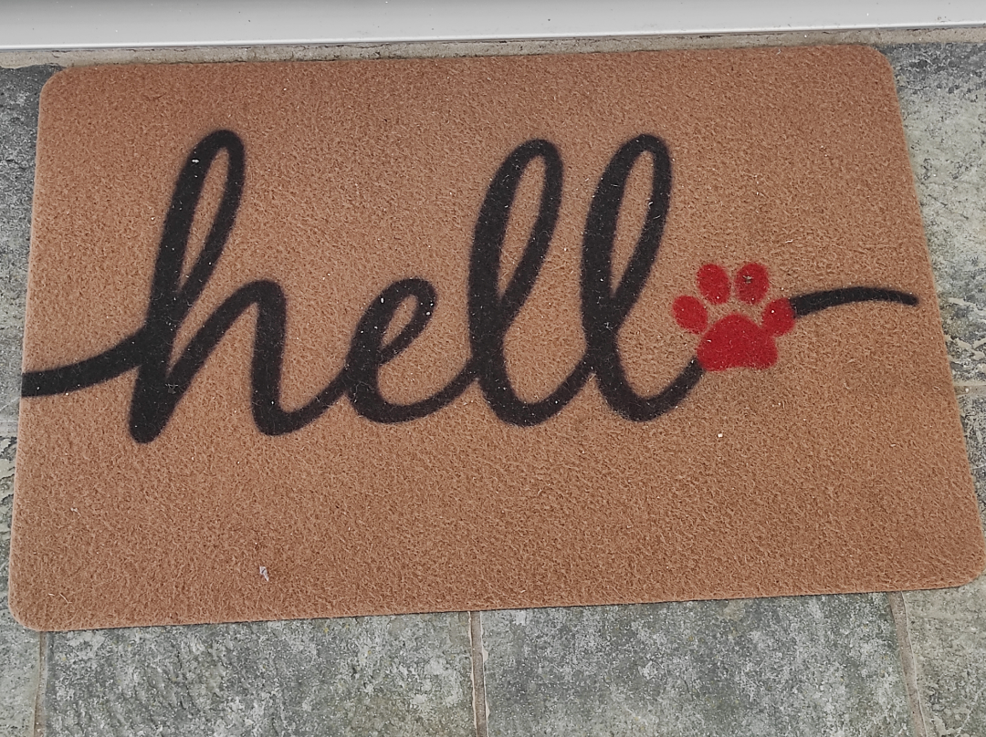 A brown doormat with the word "hello" written in black cursive, but the letter "o" is replaced by a red paw print. The mat is placed on a gray tiled floor.