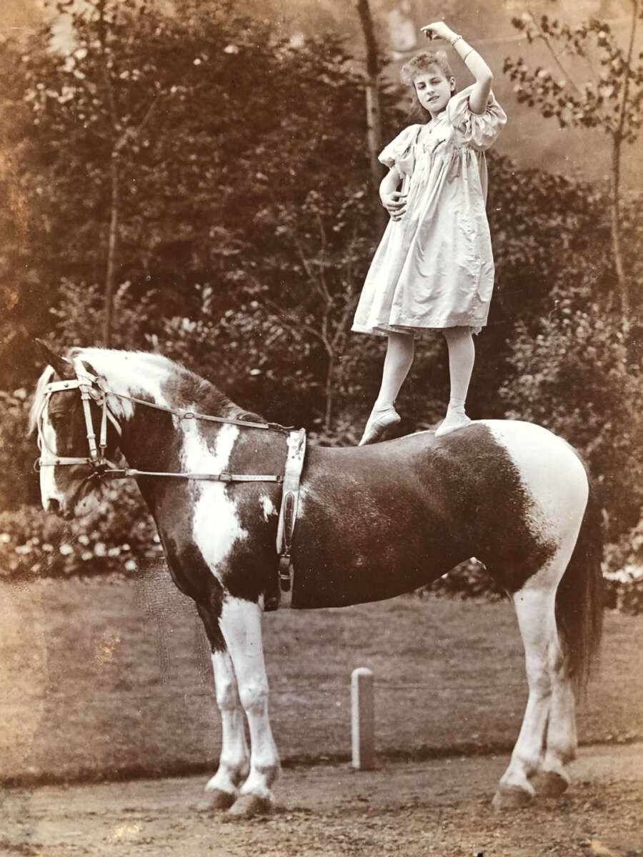 A young girl in a light dress stands on one foot on the back of a spotted horse outdoors, raising one arm gracefully, with trees and foliage in the background.