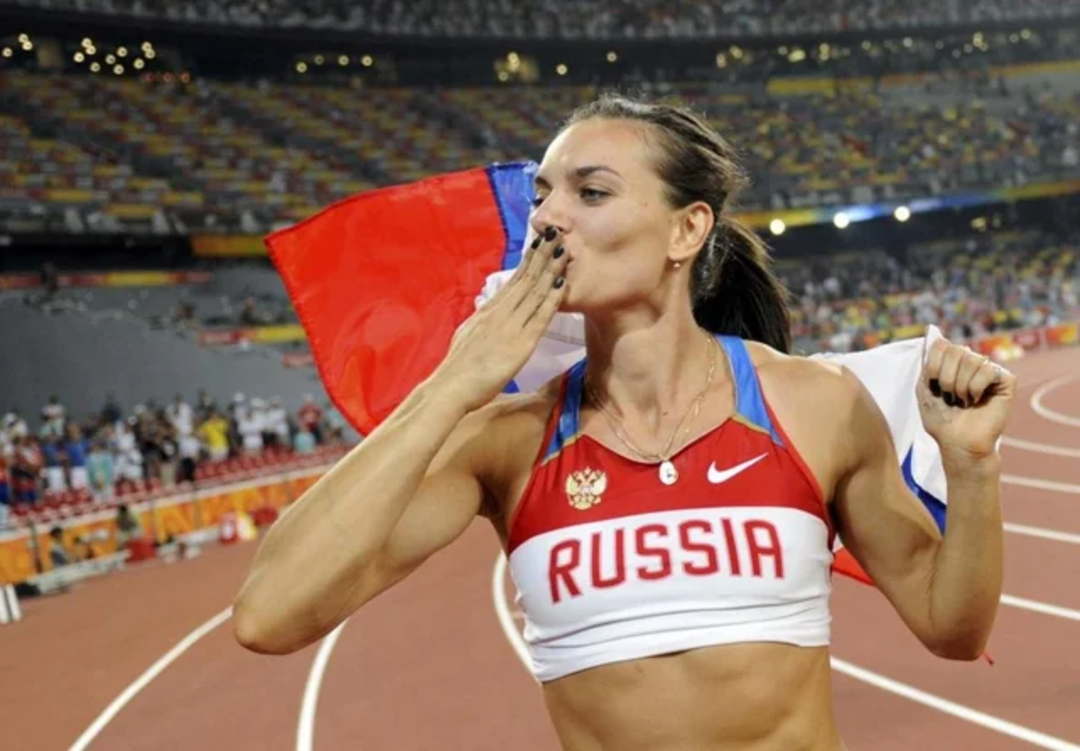 A female athlete wearing a “RUSSIA” uniform kisses her hand while holding a Russian flag over her shoulders on a track, with a stadium full of spectators in the background.